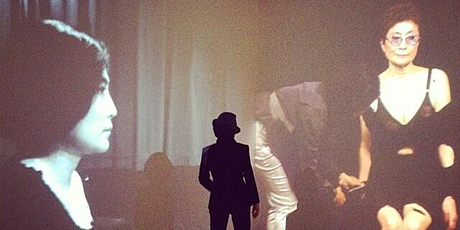 Yoko Ono stands between two screens showing her performing Cut Piece in 1965 and 2003.